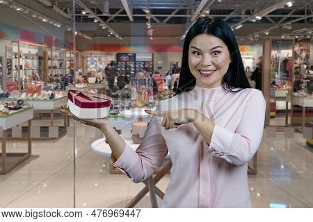 Brunette Asian Woman Pointing At Heart Shape Gift Box. Shoe Boutique Interiror In The Background.