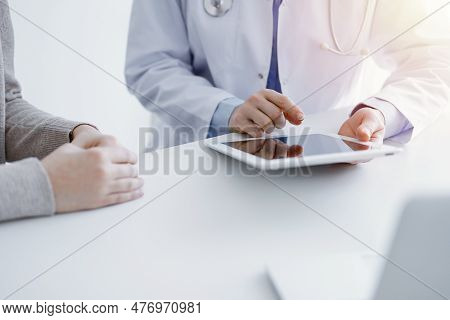 Doctor And Patient Sitting At The Table In Clinic. The Focus Is On Female Physicians Hands Using Tab