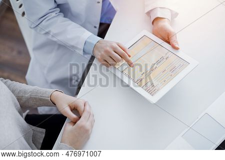 Doctor And Patient Sitting At The Table In Clinic. The Focus Is On Female Physicians Hands Using Tab