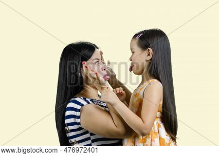 Side View Of Woman And Little Girl Are Making Faces To Each Other. Isolated On Pastel Yellow Backgro