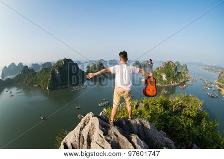 A young traveler man sit on the top of mountain with his guitar in Halong bay and enjoy the beauty of seascape.