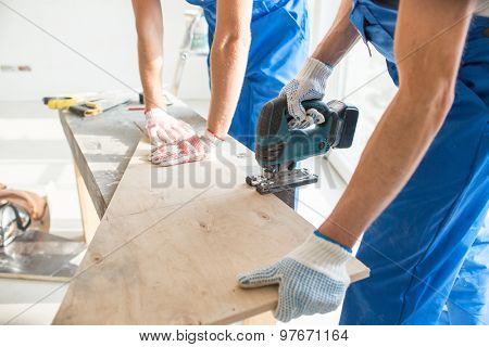 carpentry, building, teamwork and people concept - close up of builders with electric saw sawing board or wooden panel indoors