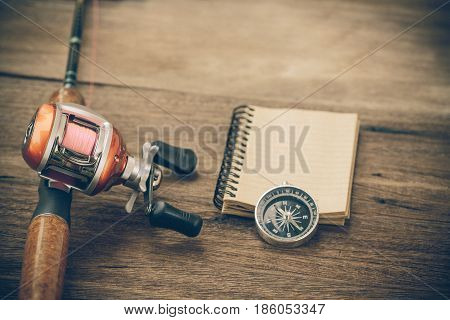 Fishing Tackle - Baitcasting Reel, Book And Compass On  Wooden Background