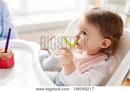childhood and people concept - little baby drinking from spout cup sitting in highchair at home