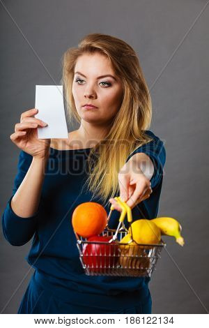 Worried woman holding shopping basket with fruits looking at bill receipt being scared of huge prices