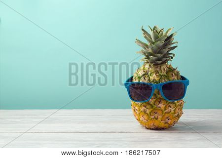 Pineapple with sunglasses on wooden table over mint background. Tropical summer vacation and beach party concept.