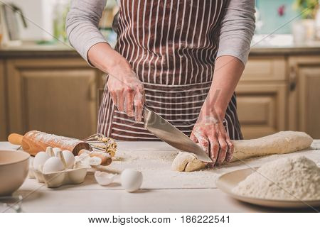 Close-up view of two woman's hands cut knife dough. A woman in a striped apron is cooking in the kitchen