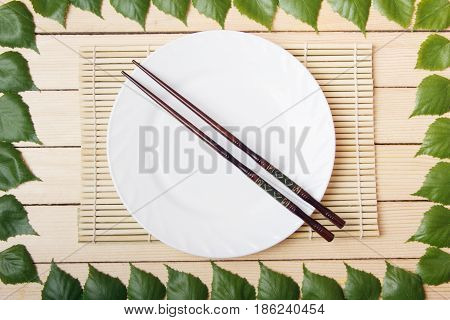 Empty plate on a bamboo mat with chopsticks top view on a wooden background framed with leaves of a tree