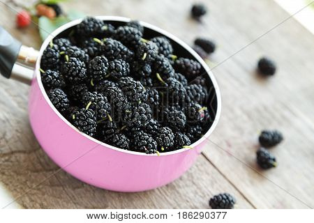 black mulberry berries in a pink pot on a wooden table. in the foreground are mulberry berries and sprig with ripened berries.