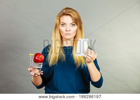 Worried woman holding shopping basket with fruits looking at bill receipt being scared of huge prices