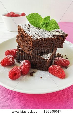 Sliced black bean brownie cake decorated with powdered sugar and mint and raspberry on white plate pink cloth napkin on white wooden table selective focus vertical close up