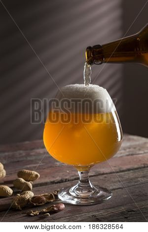 Detail of a glass of cold unfiltered pale beer being poured from a bottle with some peanuts next to the glass. Selective focus