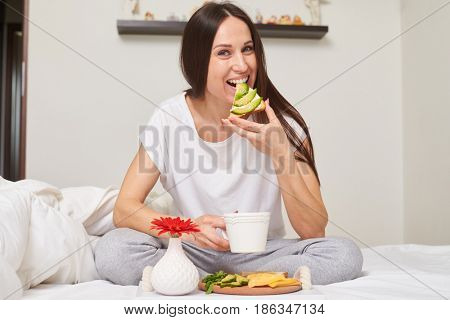 Mid shot of woman in pajamas drinking tea and eating sandwich with cheese, avocado sitting in the bed with copy space