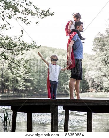 Happy loving family dad with two daughters plays a super hero in the open air near the river on the wooden pierthe concept of a super hero and a child's dreamsfather's Day