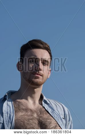 guy. Handsome young sexy caucasian model or man with bearded face and stylish hair haircut posing in unbutton shirt with naked hairy chest on sunny summer day on blue sky background. Male beauty