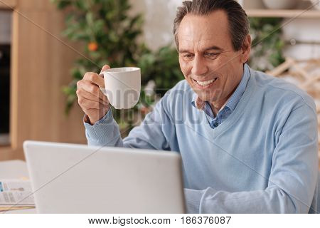 Reading morning news. Smiling glad senior man sitting in the kitchen while enjoying cup of coffee and using laptop