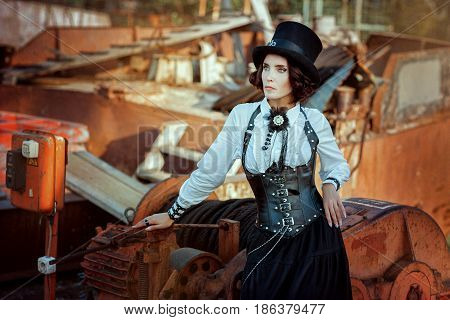 Woman dressed in a suit and a hat in a steampunk style stands next to the machines.