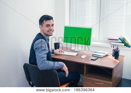 Happy office worker smiling and looking at the camera. He sits at computer with Chromakey. Office concept with working environment. Copyspace on screen