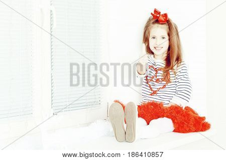 Beautiful little girl a bright red bow on head and red skirt sitting by the window. She holds a thumb up. Creative toning of a photograph.
