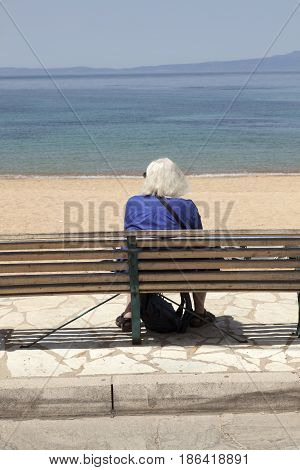 elderly lady sits alone on beach bench and looks at the sea in greek town of stoupa