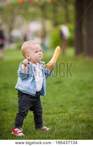 Cute little toddler boy with banana walking in the park on a summer day. First baby's steps. Outdoor. Child taking first steps on the green grass.