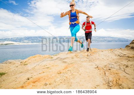 Couple runners running with backpacks on rocky trail at seaside and mountains. Young woman and man trail running on mountain path looking at beautiful inspirational landscape view.