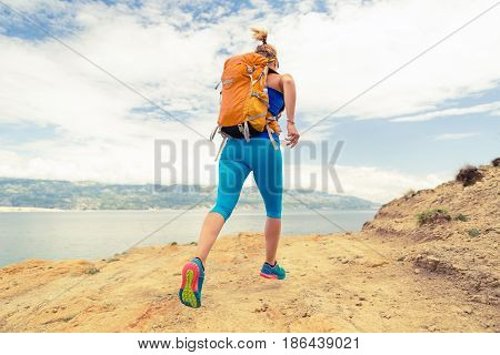 Woman running with backpack on rocky trail at seaside and mountains. Young woman training and trail running on mountain rocky dirt path looking at beautiful inspirational landscape view.