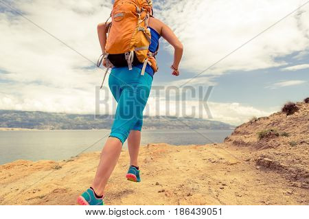 Woman running with backpack on rocky trail at seaside and mountains. Young woman training and trail running on mountain rocky dirt path looking at beautiful inspirational landscape view.