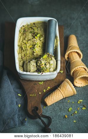 Homemade pistachio ice cream in ceramic mold with metal scooper, crashed pistachio nuts and waffle cones over dark concrete background, selective focus