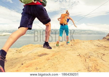 Couple runners running with backpacks on rocky trail at seaside and mountains. Young woman and man trail running on mountain path looking at beautiful inspirational landscape view.