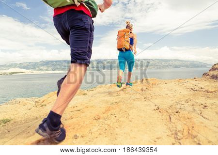 Couple runners running with backpacks on rocky trail at seaside and mountains. Young woman and man trail running on mountain path looking at beautiful inspirational landscape view.