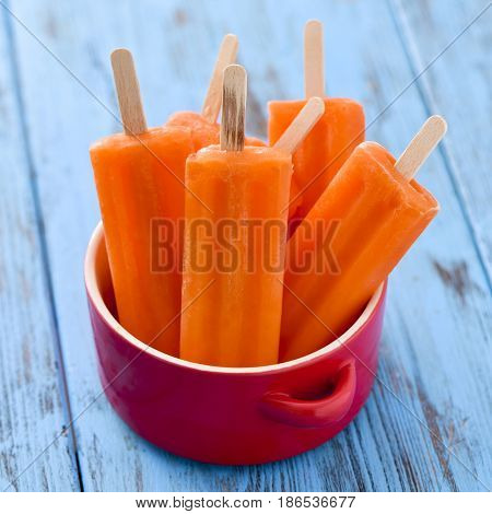 some refreshing orange flavored ice pops in a red ceramic bowl, on a blue rustic wooden table