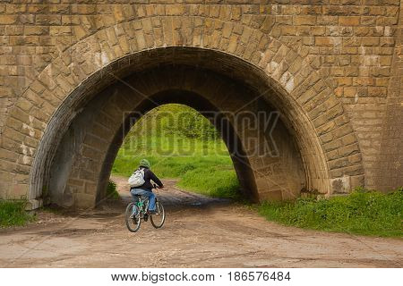 A man on a bicycle rides in a tunnel under a railway. The old tunnel is made of stone.