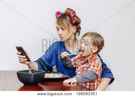 Busy tired white Caucasian young woman mother housewife with hair-curlers in hair looking on phone surfing Internet funny child son boy sitting eating meal lunch white background