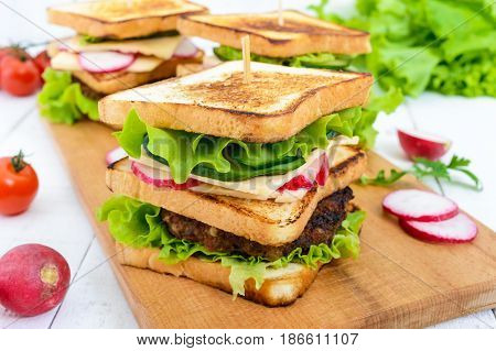Multilayered sandwiches with a juicy cutlet cheese radish cucumber lettuce arugula on a cutting board on a white wooden background.