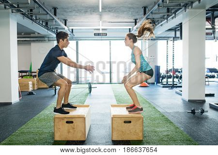 Beautiful young fit couple exercising in modern crossfit gym, doing box jumps.