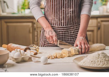 Close-up view of two woman's hands cut knife dough. A woman in a striped apron is cooking in the kitchen