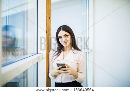Young Woman Busy Using Her Mobile Phone While Standing In Front Of Large Glass Windows