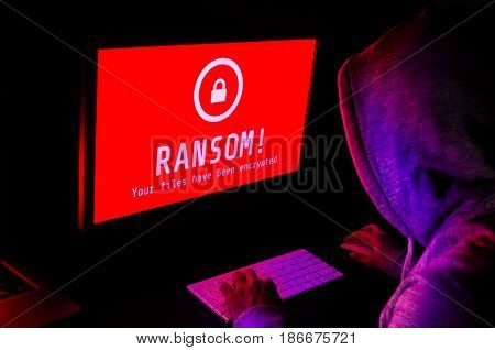 Computer screen with ransomware attack alert in red and a hacker man keying on keyboard in a dark room ideal for online security failure and digital crime