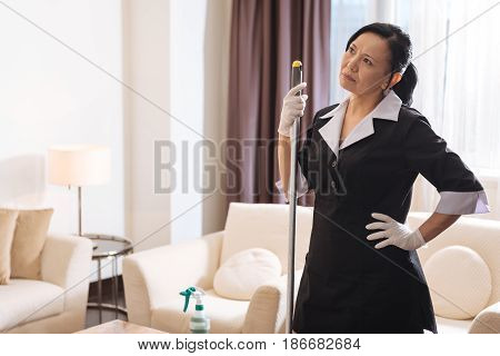 Have I done everything. Nice thoughtful pleasant woman holding a mop and thinking about her work while standing in the middle of the hotel room