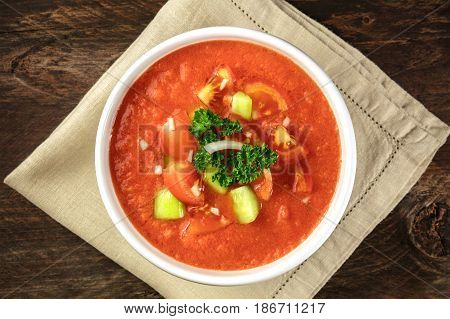 A bowl of gazpacho soup with slices of tomato, cucumber, and onion, with parsley leaves, shot from above on a dark rustic texture with a place for text