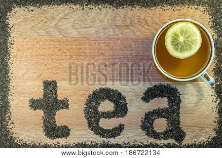 On the Board is a mug with black tea. In a mug slice of yellow lemon. On the brown Board from the scattered tea leaves of black tea small leaf lined with the letters of the word tea.