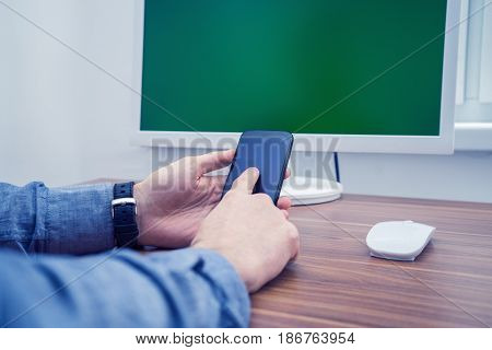 Man in office typing on telephone. computer with Chromakey is on background. Office concept with working environment. Copyspace on screen