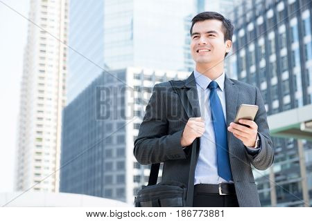 Smiling businessman using mobile phone while carrying bag and walking in the city - business travel and mobile roaming concepts