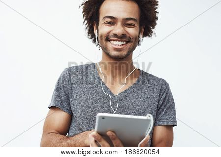 Cheerful african man in headphones smiling holding tablet looking at camera. White background. Copy space.