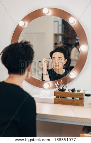 Attractive woman primp at the mirror with lamps in the beauty studio. A happy satisfied customer of make-up and hairstyle service.