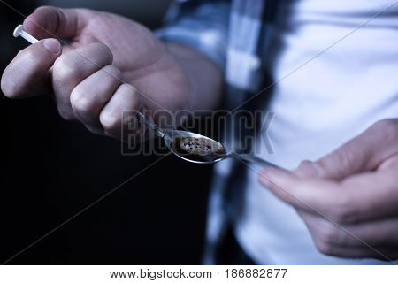 Not a single day without drugs. Skilled young obsessed freak standing in the dark place while preparing heroin dose and holding spoon and syringe