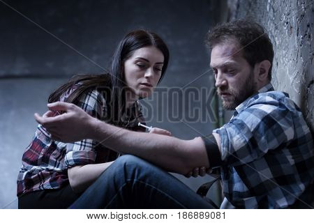 Indifference to any kind of pain. Lonely homeless obsessed couple sitting on the ground in the darkness while sharing drug dose and making syringe injection of heroin