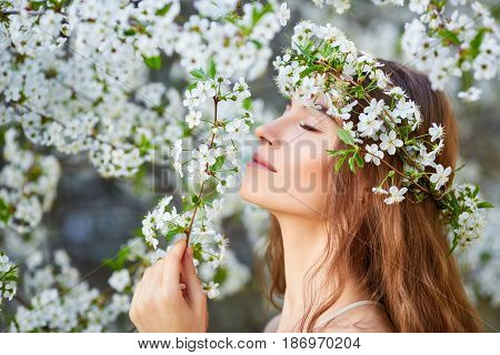 Young Beautiful Woman In Circlet Of Flowers Enjoying Smell Of Blooming Tree On A Sunny Day. Close Ey
