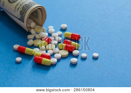 Scattered Pharmaceutical Medicine Pills, Tablets And Capsules On Dollar Money Isolated On Blue Backg
